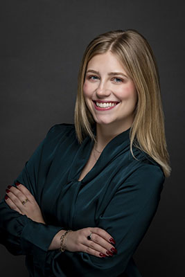 Kinsey Turk, ACAS. A confident professional woman is posed against a dark, neutral background. She is smiling warmly, showing approachability and friendliness. Her straight, blonde hair frames her face neatly, and she wears a dark teal blouse with a soft bow detail at the neckline. Her arms are crossed casually, highlighting her polished jewelry and manicured nails in a deep red shade. The overall impression is one of professionalism, confidence, and warmth.