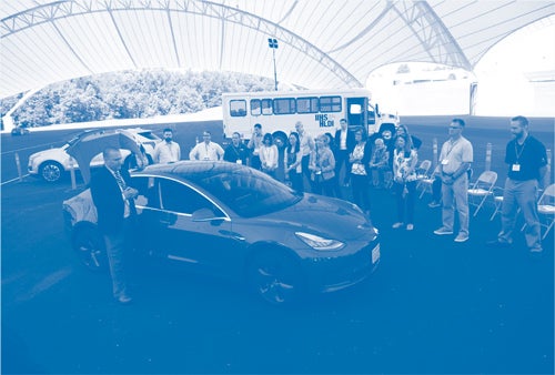 Seminar attendees gather around a Tesla Model 3 while hearing about the hands-on testing opportunities available to them on IIHS/HDLI’s test track.