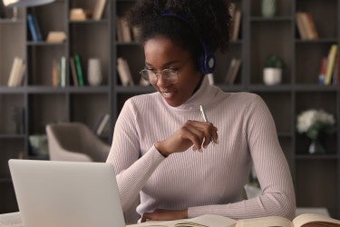 Black woman with glasses, headphones and holding pen, studying in front of laptop and open books