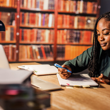 Woman studying at a library table with a laptop, notebook, and pen.