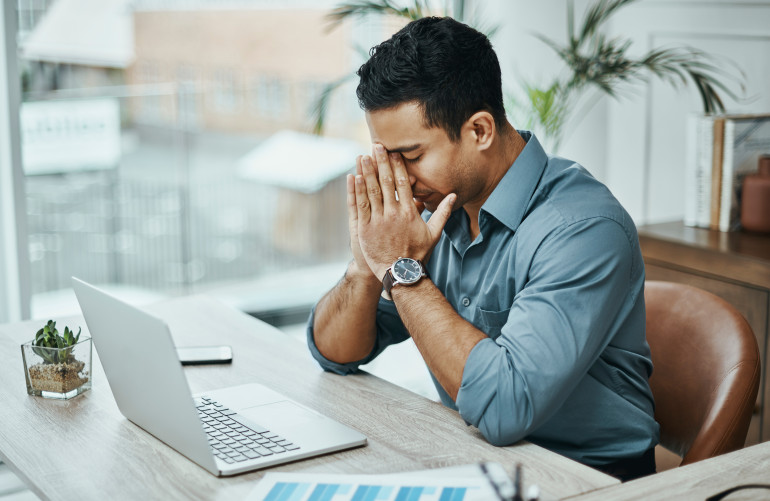 Man sitting at a desk with his head in his hands beside a laptop.
