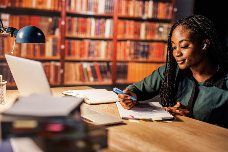 Woman studying at a library table with a laptop, notebook, and pen.